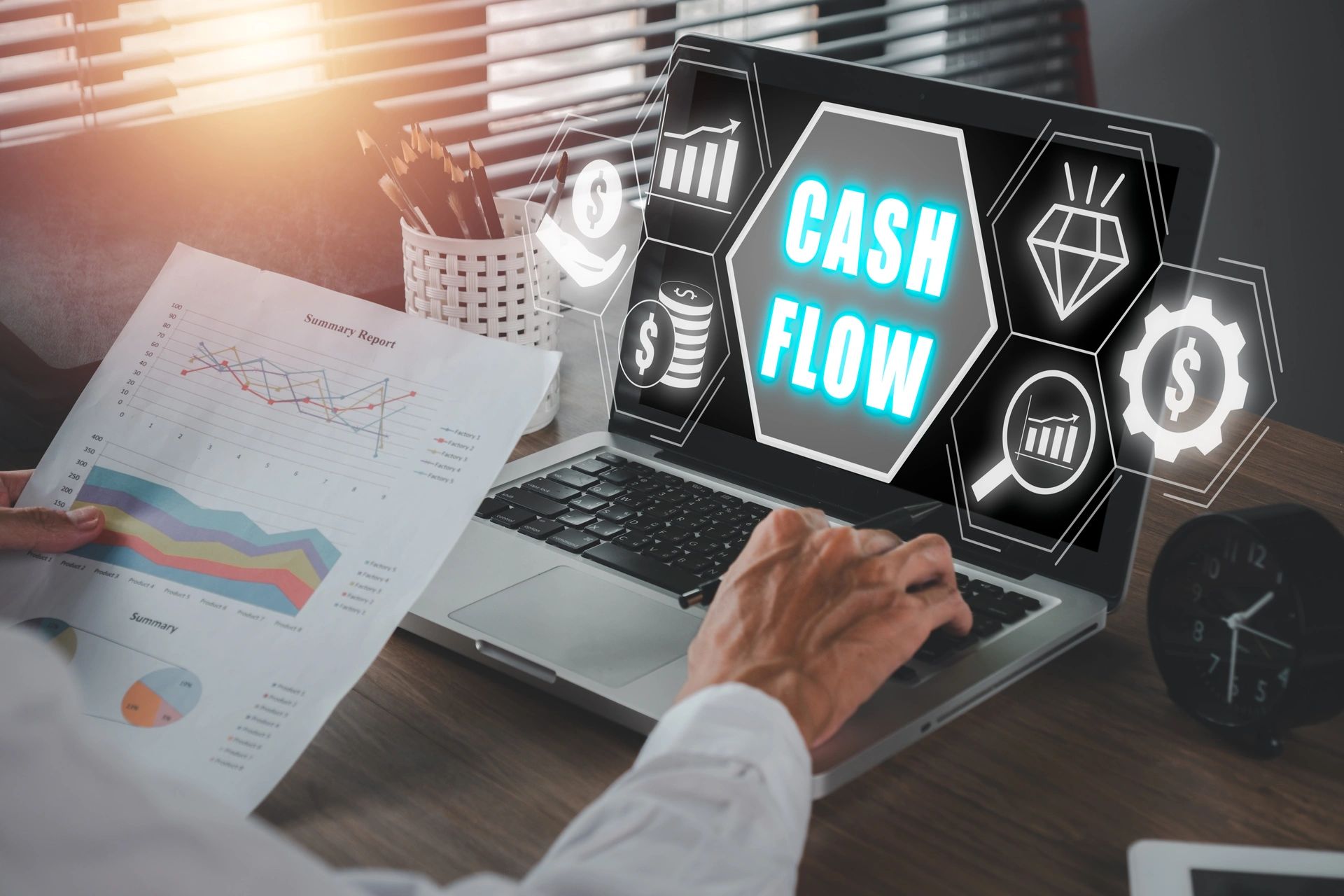 In the image, a person is working on a laptop that displays the words "CASH FLOW" prominently on the screen, along with various financial icons and graphics. The person is holding a summary report with graphs and charts, indicating financial analysis and data review. The workspace is well-organized, with pencils in a holder, and sunlight streaming through blinds, adding a productive ambiance. The setting suggests a focus on financial management and planning.