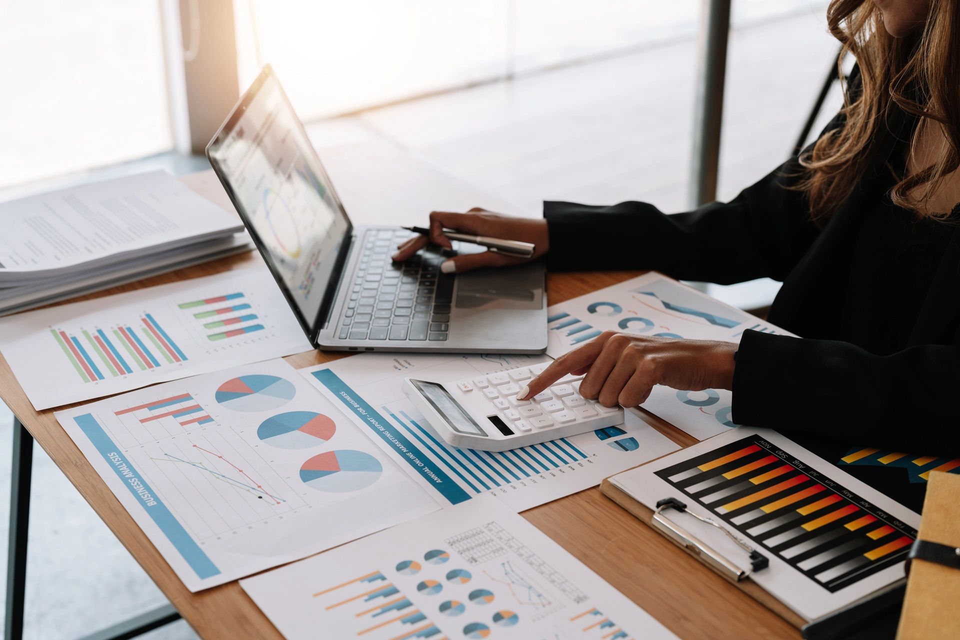 A close-up view of a bookkeeper or financial inspector's hands working on a report, using a calculator and a laptop. The desk is covered with various financial documents and charts, illustrating the concepts of bookkeeping and cash flow management.